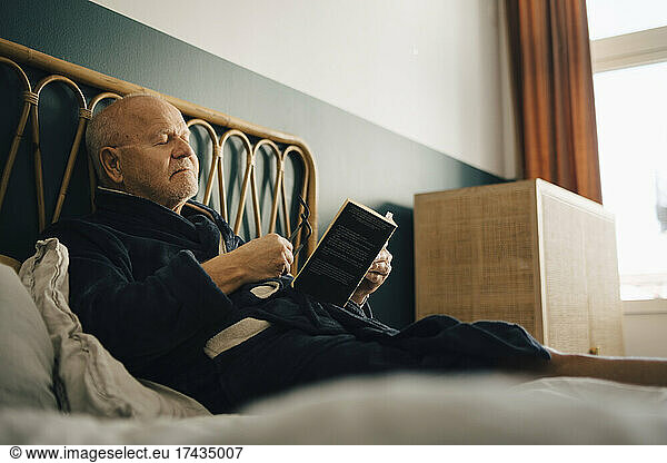 Senior man with eyes closed holding book while sitting on bed