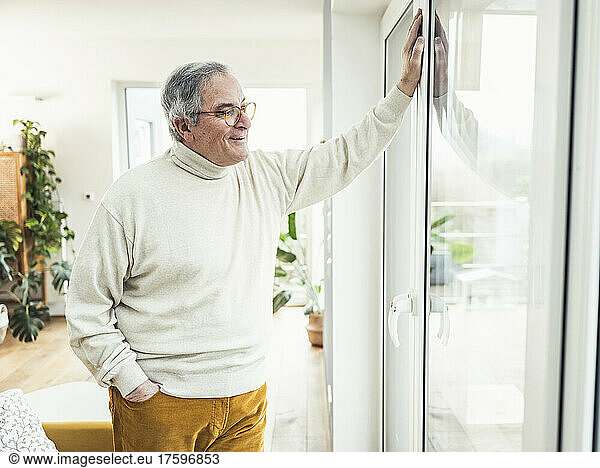 Senior man with hand in pocket looking through window at home