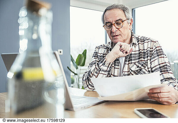 Senior man with hand on chin reading financial bill sitting at table