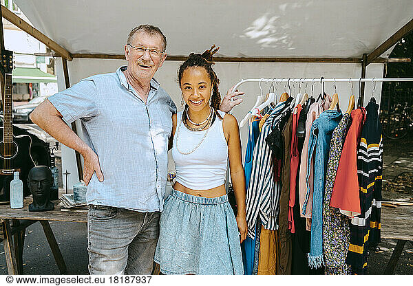 Senior man with hand on hip standing by female partner near clothes rack at flea market