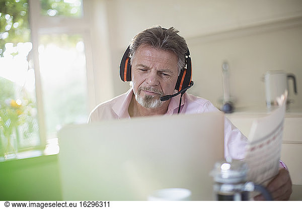 Senior man with headphones and newspaper working at laptop in kitchen