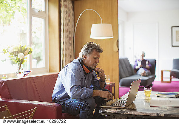 Senior man with headphones working at laptop on living room sofa