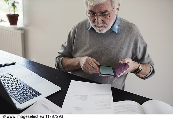 Senior man with laptop holding mobile phone while reading bills at table