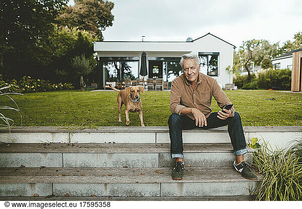 Senior man with smart phone sitting by dog on steps at backyard