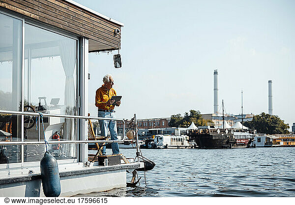 Senior man with tablet PC standing by railing at houseboat on sunny day