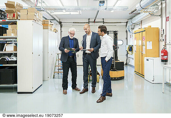 Senior manager with colleagues discussing over machine part while standing at illuminated factory