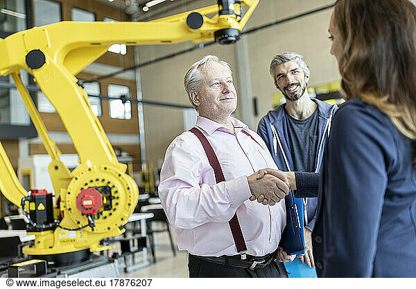 Senior technician shaking hands with female colleague in factory with industrial robots
