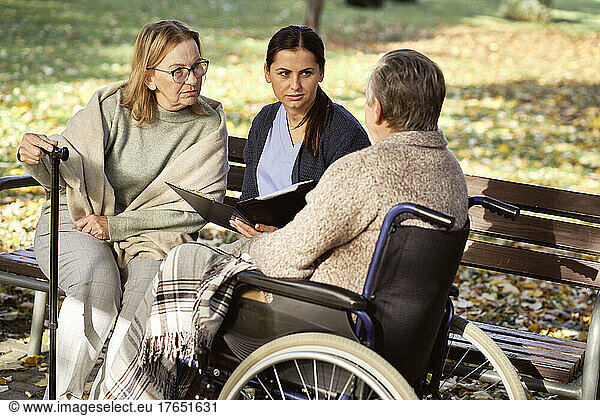 Senior woman and nurse with documents looking at disabled man sitting in wheelchair at park