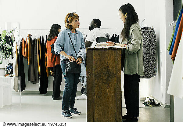 Senior woman discussing with female sales staff while standing at checkout counter in clothes store