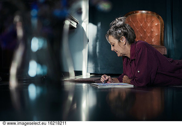Senior woman drawing in book while lying on floor at home