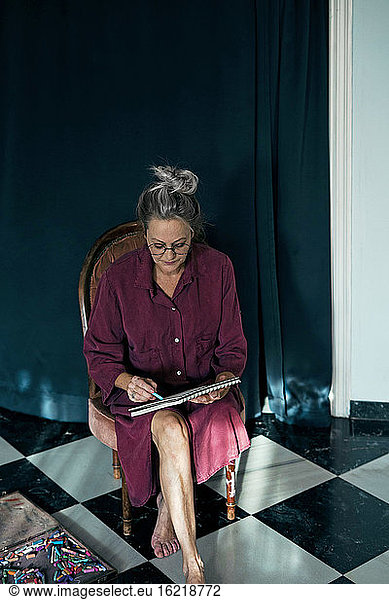 Senior woman drawing in book while sitting on chair against curtain in room