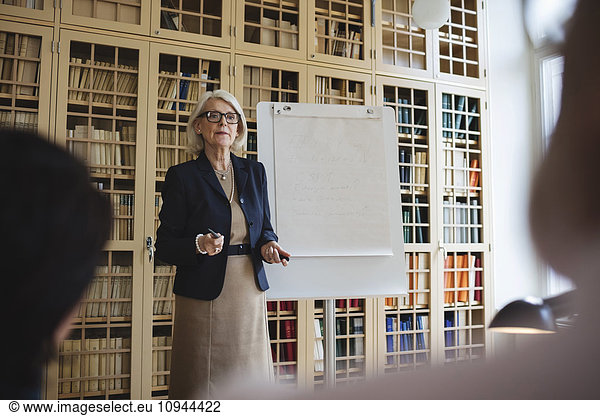 Senior woman giving presentation while standing against bookshelf
