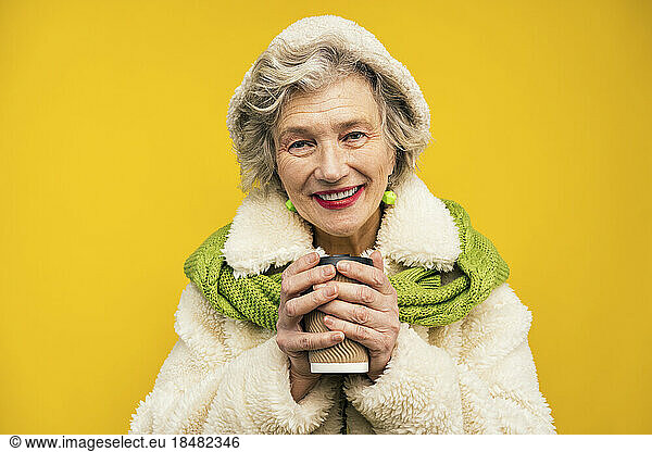Senior woman holding disposable coffee cup against yellow background