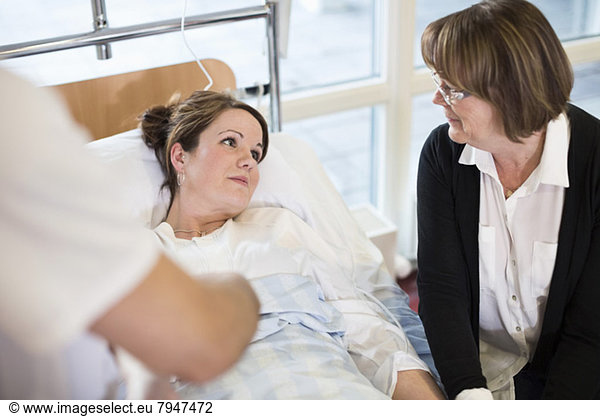 Senior woman looking at daughter lying on bed in hospital ward