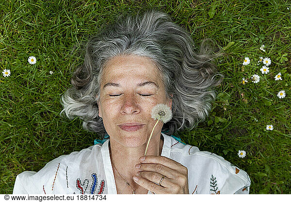 Senior woman lying on green grass holding dandelion blowball