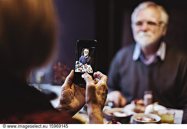 Senior woman photographing man through smart phone while having breakfast at table in hotel