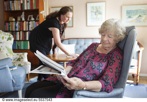 Senior woman reading newspaper while granddaughter cleaning in background