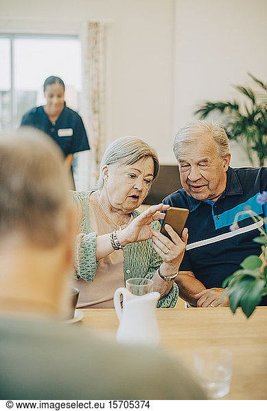 Senior woman sharing smart phone with man while sitting at dining table in retirement home