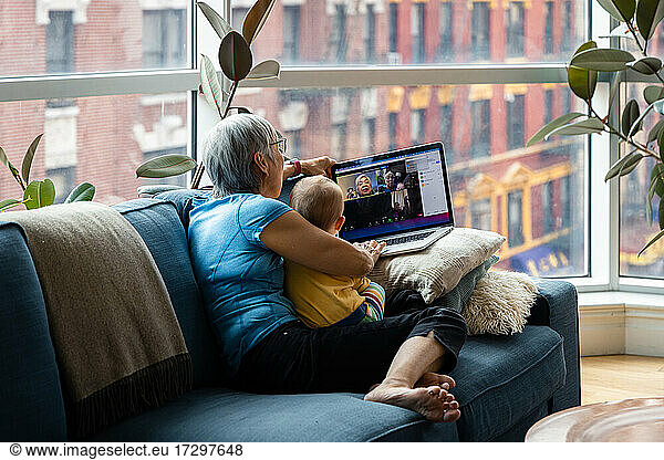 Senior woman sitting with granddaughter doing video call through laptop in living room at home