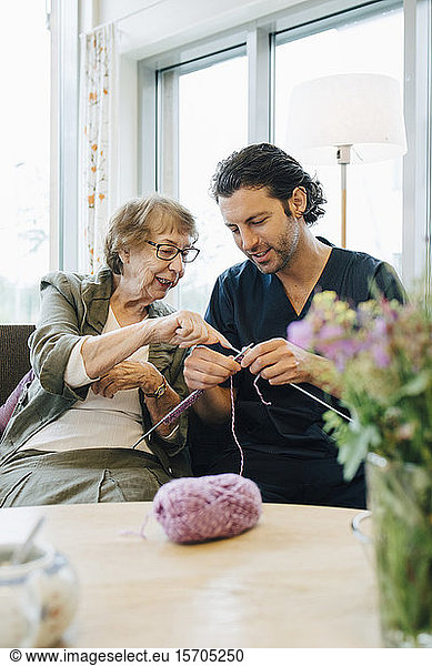 Senior woman teaching knitting to male nurse while sitting on sofa at elderly care home
