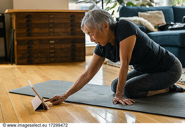 Senior woman using digital tablet while sitting on exercise mat in living room