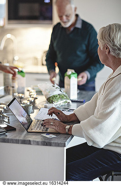 Senior woman using laptop while male partner standing by kitchen island