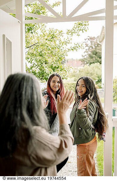 Senior woman waving hand at daughter and granddaughter from entrance of house