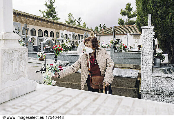 Senior woman wearing face mask putting flowers on grave in cemetery
