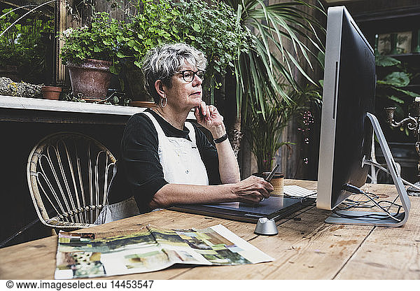 Senior woman wearing glasses  black top and white apron sitting at a wooden table  working on desktop computer.
