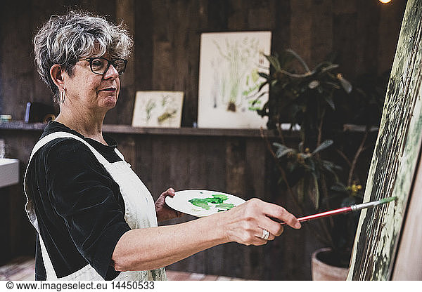 Senior woman wearing glasses  black top and white apron standing in studio  working on painting of trees in forest.