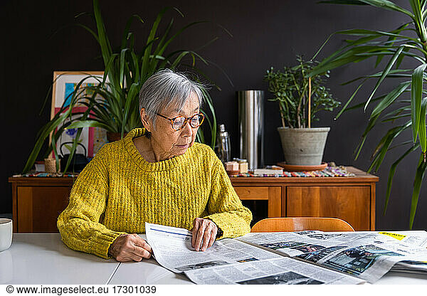 Senior woman wearing yellow sweater reading newspaper at home