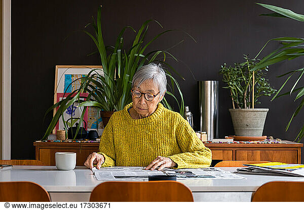 Senior woman wearing yellow sweater reading newspaper while sitting at dining table