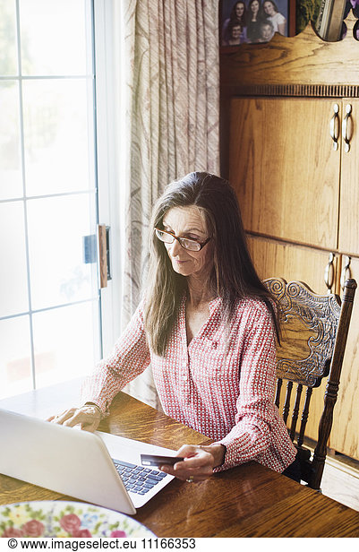 Senior woman with long brown hair sitting at a table  using a laptop computer  wearing reading glasses.