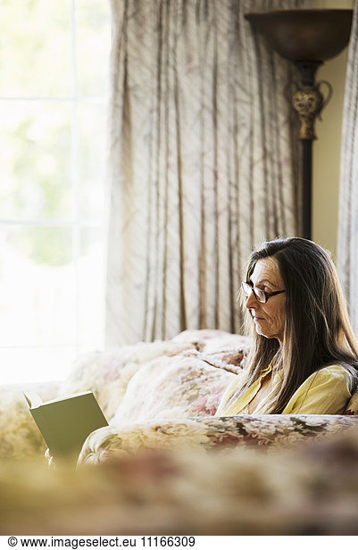 Senior woman with long brown hair sitting on a sofa  reading a book  wearing reading glasses.