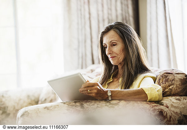Senior woman with long brown hair sitting on a sofa  using a digital tablet.