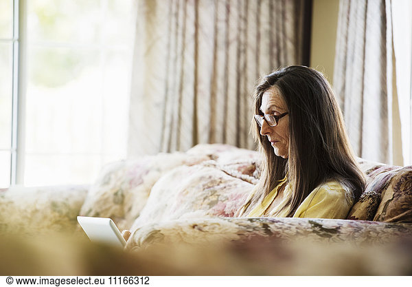 Senior woman with long brown hair sitting on a sofa  using a laptop computer  wearing reading glasses.