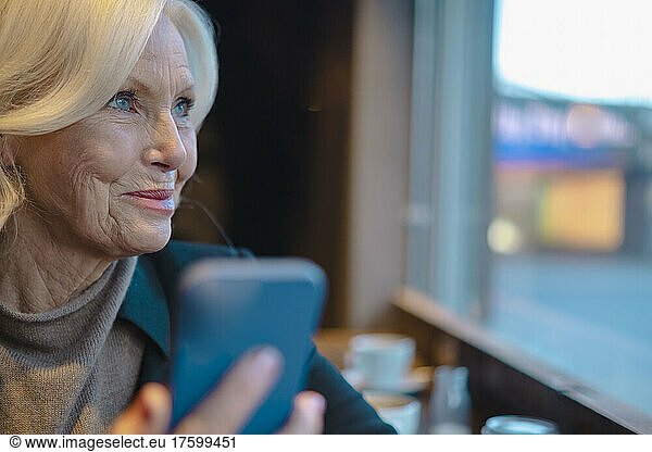 Senior woman with smart phone looking out through window