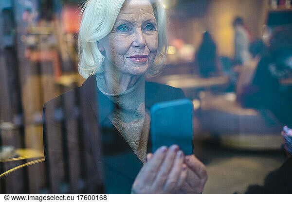 Senior woman with smart phone seen through window
