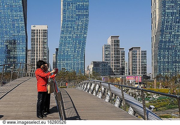 Seoul  Südkorea  Asien - Menschen auf einer Brücke vor dem Stadtbild von New Songdo City im Central Park und dem internationalen Geschäftsviertel (SIBD) mit modernen Wohntürmen. Bei dem neuen Stadtteil handelt es sich um eine intelligente Stadt oder allgegenwärtige Stadt   die von Grund auf auf 1.500 Hektar zurückgewonnenem Land entlang der Uferpromenade von Incheon  40 Kilometer südwestlich des Stadtzentrums von Seoul  errichtet wurde. Zusammen mit Yeongjong und Cheongna ist es Teil der Freien Wirtschaftszone Incheon.