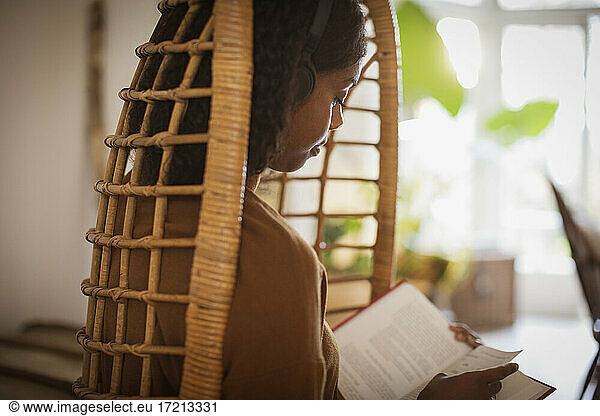Serene junge Frau mit Kopfhörer lesen Buch in Rattan Stuhl