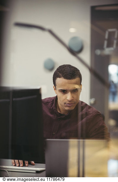 Serious businessman using laptop at desk seen through glass in office