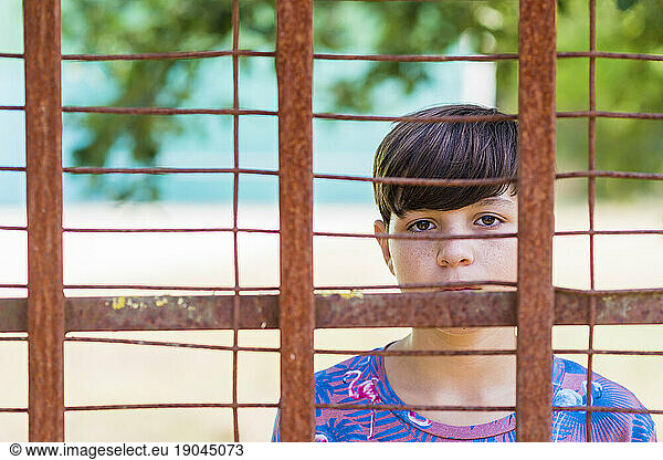 serious teen in casual wear behind metallic fence outdoors