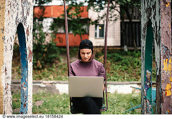 Serious young female student using laptop sitting on a swing in summer