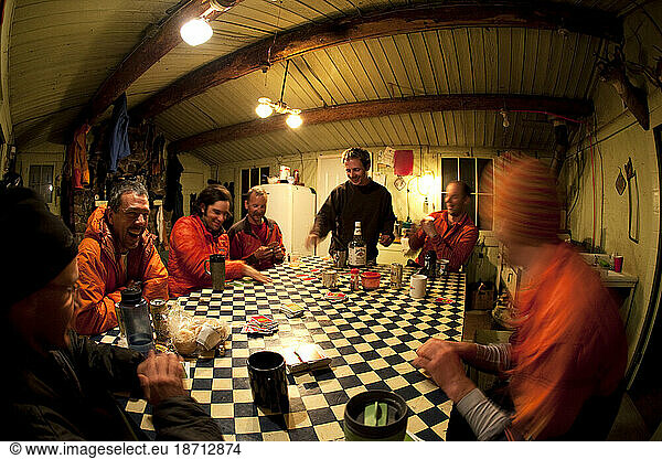 Seven men in ski clothes hanging out  drinking  playing cards and laughing in a rustic cabin at night.