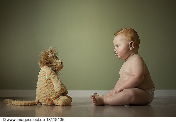 Shirtless baby boy looking at stuffed toy while sitting on floor against wall at home