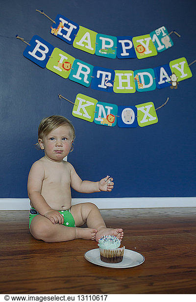 Shirtless baby boy sitting on floor against birthday banner hanging on wall at home