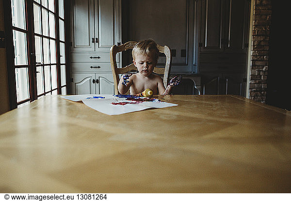 Shirtless baby boy with dirty hands sitting by wooden table at home
