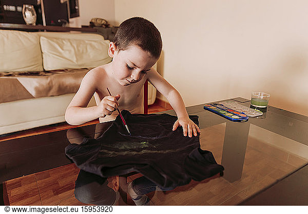 Shirtless boy drawing on black t-shirt at table in living room