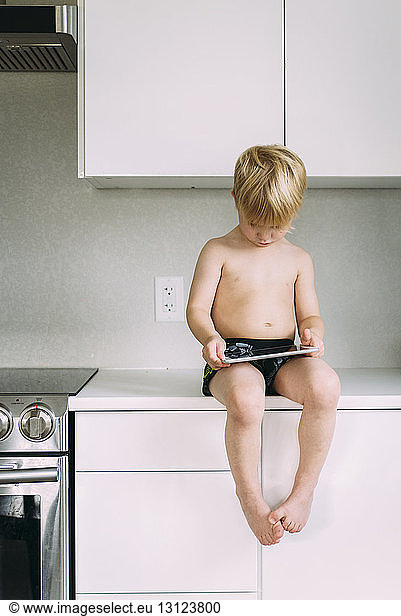 Shirtless boy using tablet computer while sitting on kitchen counter at home