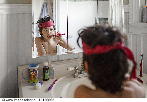 Shirtless boy with red textile tied on head brushing teeth while looking at his reflection in mirror